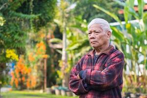 Porträt von ein Alten asiatisch Mann mit grau Haar, Waffen gekreuzt und suchen Weg während Stehen im ein Garten. Konzept von alt Menschen und Gesundheitswesen foto