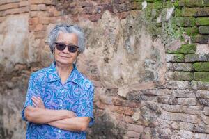 ein Porträt von ein Alten asiatisch Frau tragen Sonnenbrille, Waffen gekreuzt, lächelnd, und suchen beim das Kamera während Stehen mit alt Backstein Mauer Hintergrund. Konzept von alt Menschen und Entspannung foto