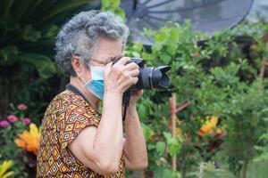 Senior Frau tragen ein Gesicht Maske und Schießen ein Foto durch das Digital Kamera während Stehen im ein Garten. Maske zum schützen Virus, Coronavirus. Neu normal. Konzept von alt Menschen und Fotografie