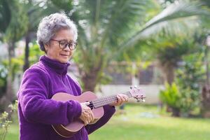 Porträt von ein Alten asiatisch Frau spielen das Ukulele während Stehen im das Garten. Raum zum Text. Konzept von alt Menschen und Entspannung foto