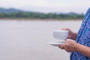 Nahansicht von Hände Senior Frau halten ein Weiß Kaffee Tasse während Stehen beim das Seite das Fluss. Morgen Kaffee mit ein gemütlich Atmosphäre. Raum von Text. Konzept von alt Menschen und Entspannung foto