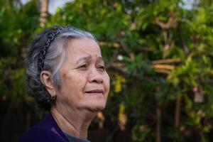 Porträt von ein schön Senior Frau mit kurz grau Haar lächelnd und suchen Weg während Stehen im ein Garten. Raum zum Text. Konzept von alt Menschen und Entspannung. Seite Aussicht foto