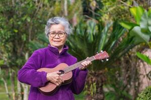 heiter Alten asiatisch Frau mit kurz grau Haar tragen Brille und spielen das Ukulele während Stehen im ein Garten. Konzept von alt Menschen und Entspannung foto