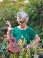 Porträt von ein Alten asiatisch Frau mit kurz grau Haar halten ein Ukulele während Stehen im ein Garten. Konzept von alt Menschen und Entspannung foto