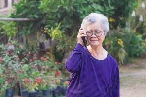 ein Porträt von ein Alten Frau mit Handy, Mobiltelefon Telefon während Stehen im ein Garten. Raum zum Text. Konzept von alt Menschen und Telekommunikation foto