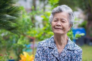 ein Porträt von ein Alten Frau lächelnd und suchen beim Kamera während Stehen im ein Garten. foto