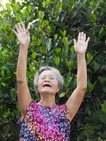 Porträt von ein Alten Frau Übungen durch gestikulieren erziehen beide Waffen, lächelnd, und suchen beim das Kamera während Stehen im ein Garten. Raum zum Text. Konzept von alt Menschen und Gesundheitswesen foto