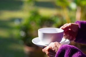 Nahansicht von Hände Senior Frau halten ein Weiß Kaffee Tasse während Stehen im das Garten. Raum zum Text. Konzept von alt Menschen und Entspannung foto