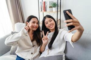 zwei Frauen sind lächelnd und nehmen ein Selfie Sitzung auf das Sofa im das Leben Zimmer beim heim. foto
