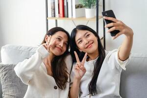 zwei Frauen sind lächelnd und nehmen ein Selfie Sitzung auf das Sofa im das Leben Zimmer beim heim. foto