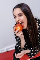 Taille oben Porträt Aussicht von das Brünette Mädchen gelehnt beim das rot Stuhl beim das Studio mit Weiß Mauer beim das Hintergrund. geduldig beißt das Apfel. gesund Zähne Konzept foto