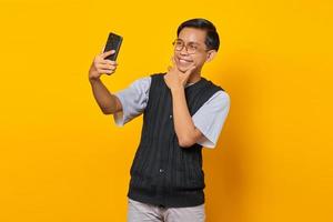 Fröhlicher asiatischer Mann mit Handy, der Selfie über gelbem Hintergrund macht foto