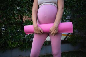 ein jung schwanger Frau hält ein Rosa Yoga Matte im ihr Hände. Nahansicht Schuss foto