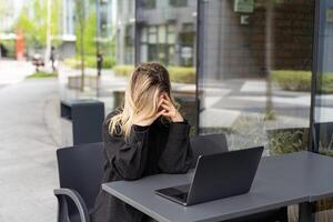 müde jung Geschäft Frau im schwarz passen mit Kopfhörer ist Sitzung mit Augen geschlossen im Vorderseite von Laptop foto