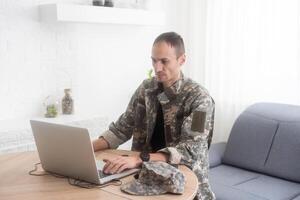 Soldat im Büro. ernst jung Militär- Mann Tippen auf Laptop während Arbeiten im Hauptquartier Gebäude. kaukasisch Mann im tarnen Uniform ist Sitzung im hell Zimmer beim Tabelle mit Notizbuch und Deckel. foto