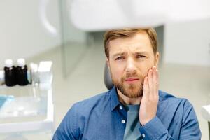 traurig Mann Sitzung im Dental Stuhl Leiden von akut Zahn Schmerz, Reiben seine Wange, schließen oben foto