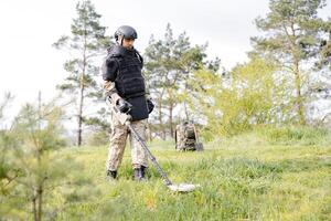 ein Mann im ein Militär- Uniform und kugelsicher Weste funktioniert im das Wald mit ein Metall Detektor. ein Minensuchboot führt aus Arbeit auf Minenräumung das Gebiet foto