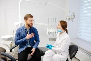 blond weiblich Zahnarzt im Dental Büro reden mit männlich geduldig und vorbereiten zum Behandlung. gut aussehend bärtig Mann im Zahnarzt Stuhl suchen beim seine Arzt mit Lächeln foto