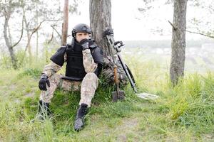 ein Mann im ein Militär- Uniform und ein kugelsicher Weste sitzt im das Wald in der Nähe von ein Metall Detektor und ein Militär- Rucksack. ein Mann pausiert im das Arbeit von Minenräumung das Gebiet foto