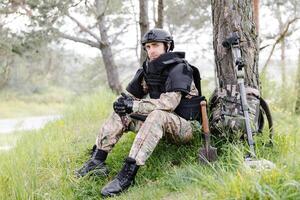ein Mann im ein Militär- Uniform und ein kugelsicher Weste sitzt im das Wald in der Nähe von ein Metall Detektor und ein Militär- Rucksack. ein Mann pausiert im das Arbeit von Minenräumung das Gebiet foto