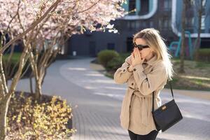 Frau allergisch Leiden von saisonal Allergie beim Frühling im blühen Garten beim Frühling. jung Frau Niesen und weht Nase mit Nasal- Taschentuch im Vorderseite von Blühen Baum. Allergie Konzept. foto