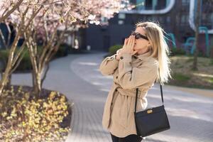 Frau allergisch Leiden von saisonal Allergie beim Frühling im blühen Garten beim Frühling. jung Frau Niesen und weht Nase mit Nasal- Taschentuch im Vorderseite von Blühen Baum. Allergie Konzept. foto