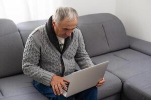 älter Mann Sitzung auf Sofa, lächelnd beim Computer Bildschirm beim Zuhause foto