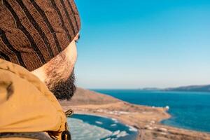 ein Mann im ein Hut und Jacke sieht aus beim das Meer Horizont und das Hügel mit vergilbt Gras. ein Mann ist Wandern entlang das hügelig Meer Ufer. russky Insel, Wladiwostok, Russland. foto