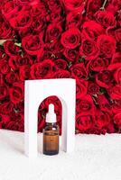 ein kosmetisch Flasche mit ein Tropfer mit ein natürlich Mittel, Öl oder Serum zum Gesicht und Körper Haut Pflege steht im ein Weiß Bogen gegen ein schick rot Rosen. foto