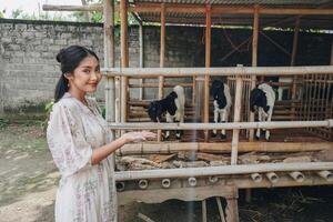 lächelnd jung asiatisch Frau Stehen im Vorderseite von traditionell Käfig gemacht von Holz und Bambus im Indonesien ländlich Bereich mit Ziege Innerhalb foto