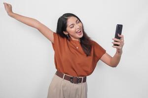 froh jung asiatisch Frau im braun Hemd tun Anruf mit glücklich erfolgreich Ausdruck und geballt Faust Geste isoliert Über Weiß Hintergrund. foto
