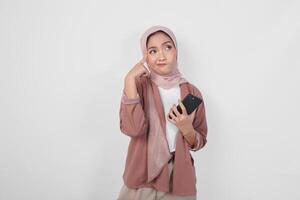nachdenklich asiatisch Muslim Frau tragen Hijab suchen beiseite während halten Smartphone isoliert Über Weiß Hintergrund. foto