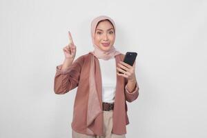 hungrig jung asiatisch Muslim Frau tragen Hijab zeigen zu das Kopieren Raum nach oben während halten Smartphone isoliert Über Weiß Hintergrund. foto