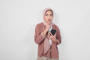 nachdenklich asiatisch Muslim Frau tragen Hijab suchen beiseite während halten Smartphone isoliert Über Weiß Hintergrund. foto