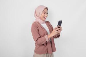 jung asiatisch Muslim Frau tragen Hijab lächelnd und suchen zu das Smartphone sie hält isoliert durch Weiß Hintergrund. foto