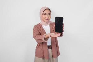 jung asiatisch Muslim Frau tragen Hijab ist lächelnd während zeigen Kopieren Raum auf ihr Smartphone Bildschirm, isoliert durch Weiß Hintergrund. foto