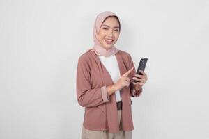 aufgeregt asiatisch Muslim Frau im Hijab zeigen zu das Smartphone sie hält Über isoliert Weiß Hintergrund. foto