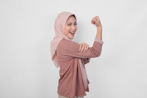aufgeregt asiatisch Muslim Frau tragen ein Hijab zeigen stark Geste durch Heben ihr Waffen und Muskeln lächelnd stolz. foto