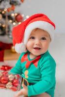 Santa's wenig Helfer Nächster zu das Weihnachten Baum mit Weihnachten Bälle im seine Hände. Porträt von Baby foto