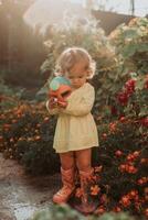 wenig Mädchen im ein Gelb Kleid und Gummi Stiefel ist Bewässerung Blumen im das Herbst Garten foto