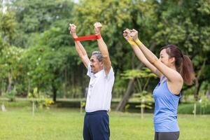 Senior asiatisch Mann und seine Tochter sind mit Sport Gummi Band zu bauen oben seine Arm Muskel Stärke im das Öffentlichkeit Park zum ältere Langlebigkeit Übung und draussen trainieren Verwendungszweck foto
