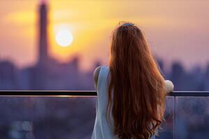 Frau suchen und genießen Sonnenuntergang Aussicht von Balkon mit das Sonne Rahmen hinter Wolkenkratzer im beschäftigt städtisch Innenstadt mit Einsamkeit zum Einsamkeit, Einsamkeit und träumend von Freiheit Lebensstil foto