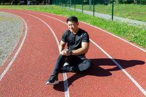 asiatisch Sportler verletzt während Joggen, Mann im Trainingsanzug Sitzung auf Stadion Laufband und halten Hand durch Bein, haben stark Schmerzen im Muskeln, Bänder, massieren foto