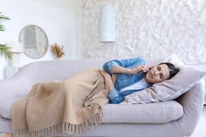 verärgert krank Frau allein beim heim, Lügen auf das Sofa unter ein Decke, Berufung das Arzt auf das Telefon foto