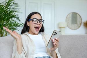 schön jung Brünette Frau tragen Brille beim Zuhause im Leben Zimmer schockiert empfangen gut Nachricht, lesen auf Telefon und Glücklich, feiern Sieg, halten Hand oben Erfolg Geste foto