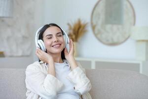 schön jung Frau ruhen Hören zu Musik- im Weiß groß Kopfhörer, Brünette Sitzung auf Sofa im Leben Zimmer mit geschlossen Augen träumend beim Zuhause foto