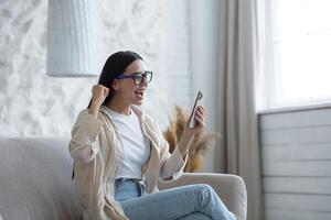 schön jung Brünette Frau tragen Brille beim Zuhause im Leben Zimmer schockiert empfangen gut Nachricht, lesen auf Telefon und Glücklich, feiern Sieg, halten Hand oben Erfolg Geste foto