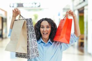 Porträt von ein glücklich Frau Käufer, spanisch Frau im ein Supermarkt mit bunt Taschen ist glücklich und suchen beim das Kamera, Innerhalb ein groß Kleidung Geschäft foto