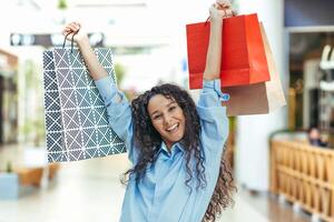 Urlaub Verkauf. schwarz Freitag. glücklich jung spanisch Frau Einkaufen im ein Supermarkt. sie hält farbig Papier Einkaufen Taschen im ihr Hände, angehoben Sie hoch, freut sich, lächelt beim das Kamera. foto