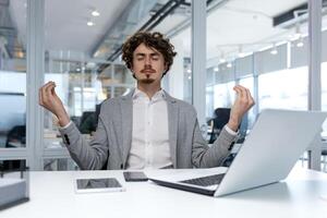 spanisch Geschäftsmann im hell Weiß Büro entspannend beim Arbeitsplatz, Mann meditieren im Lotus Position beim Tabelle mit Laptop, Financier entspannend und Atmung frisch Luft, Boss im passen mit Augen geschlossen. foto